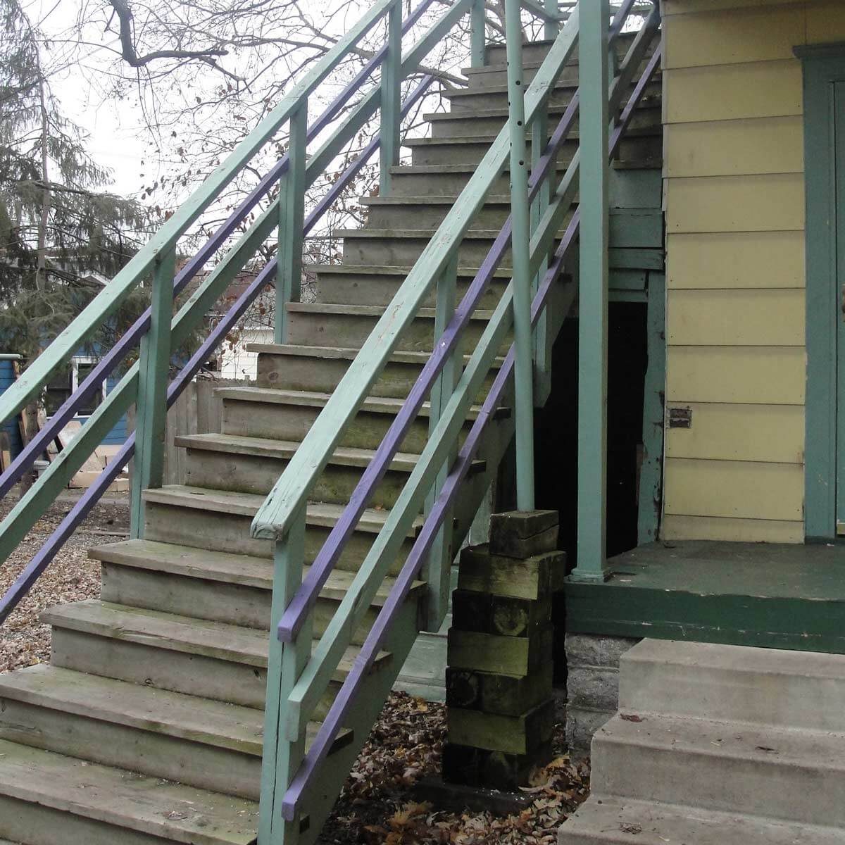 A wooden staircase with green railings and purple accents leads to a building&rsquo;s entrance, surrounded by bare trees and fallen leaves on the ground.