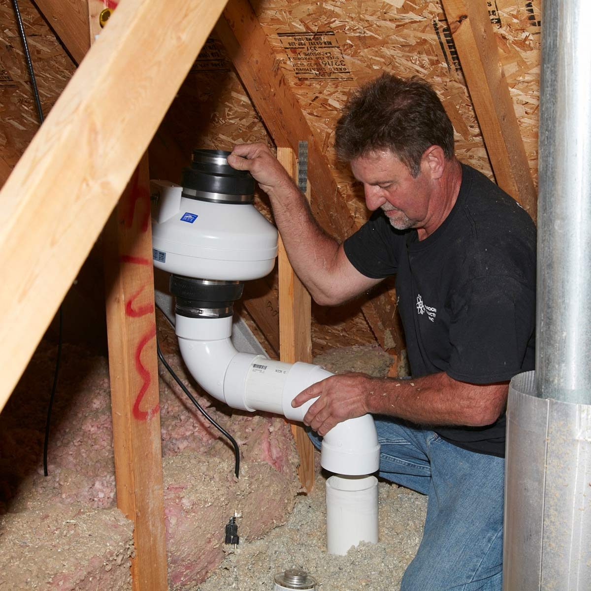 A man is installing a ventilation system component in an attic, surrounded by insulation and wooden beams, focusing on connecting white PVC pipes.