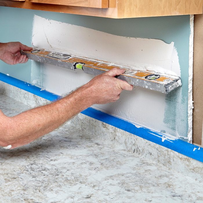 A person uses a level tool to ensure a smooth wall surface beneath kitchen cabinets, with a countertop and painter
