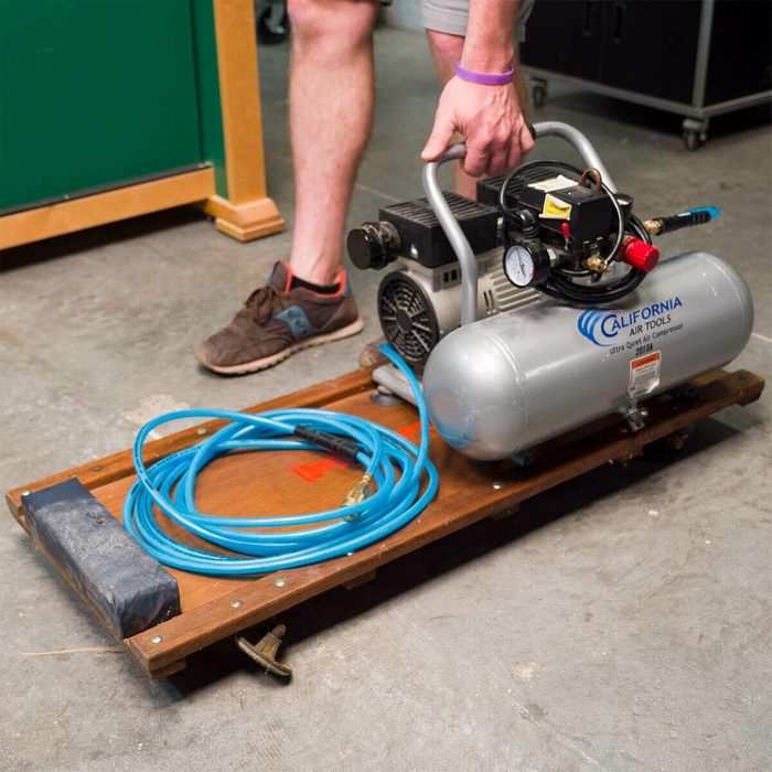 A person is lifting a portable air compressor with a blue hose coiled nearby, set on a wooden platform in a garage-like environment.
