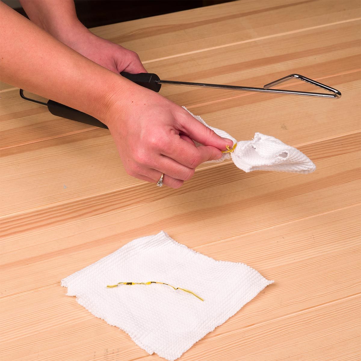 A hand uses a tool to extract a yellow item from a white cloth on a wooden surface, while another piece of cloth sits nearby.