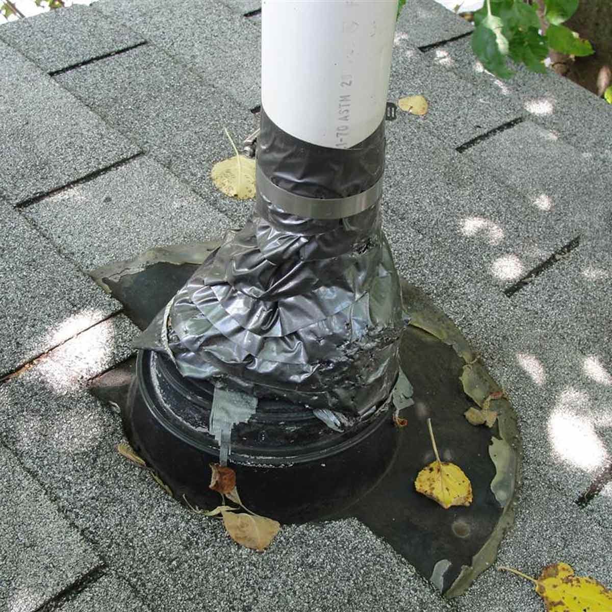 A PVC pipe is installed through a roof, surrounded by black tape for sealing. Fallen leaves are scattered on the gray asphalt shingles.