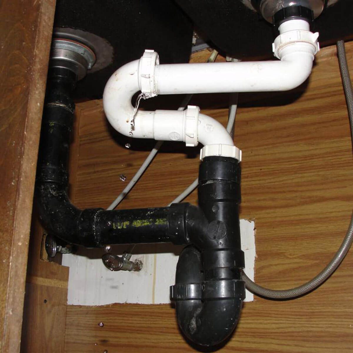 A series of pipes, including black and white PVC, are installed under a sink, with water droplets forming and trickling down.