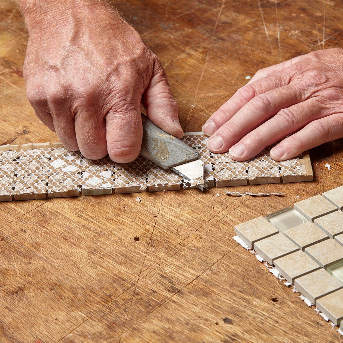 Hands are cutting mosaic tiles on a wooden surface, shaping them with a tool for a tiling project. The environment is a workspace filled with materials.