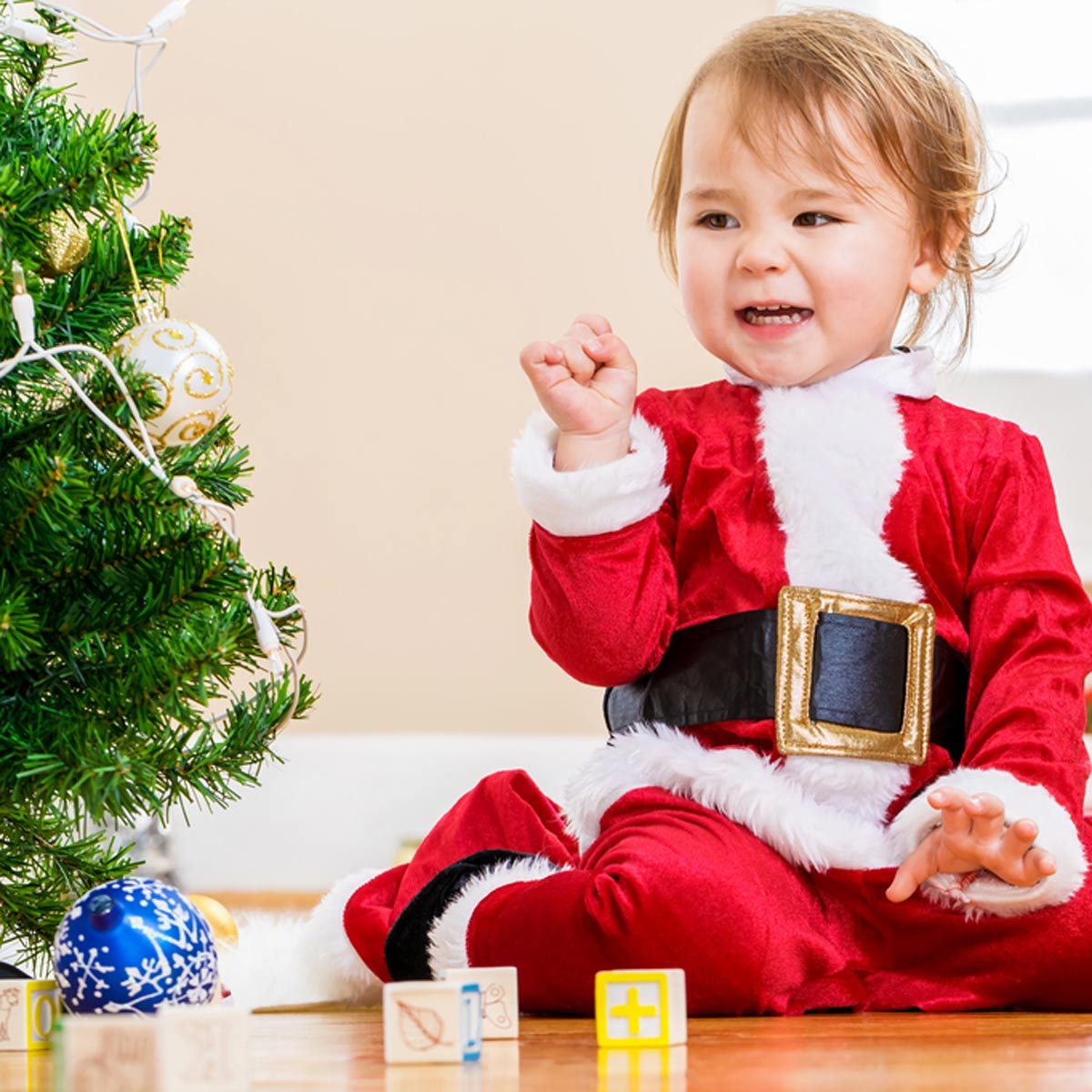 A young child in a Santa costume joyfully interacts with colorful blocks next to a decorated Christmas tree in a warmly lit room.