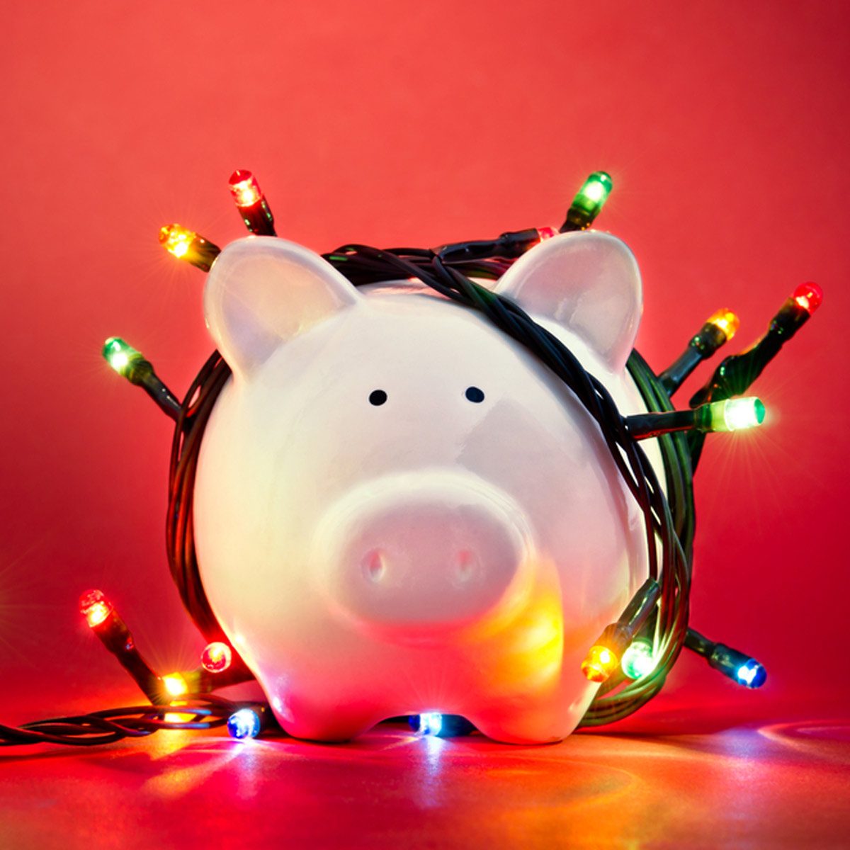A white piggy bank is wrapped in colorful Christmas lights, glowing against a red background, creating a playful holiday atmosphere.