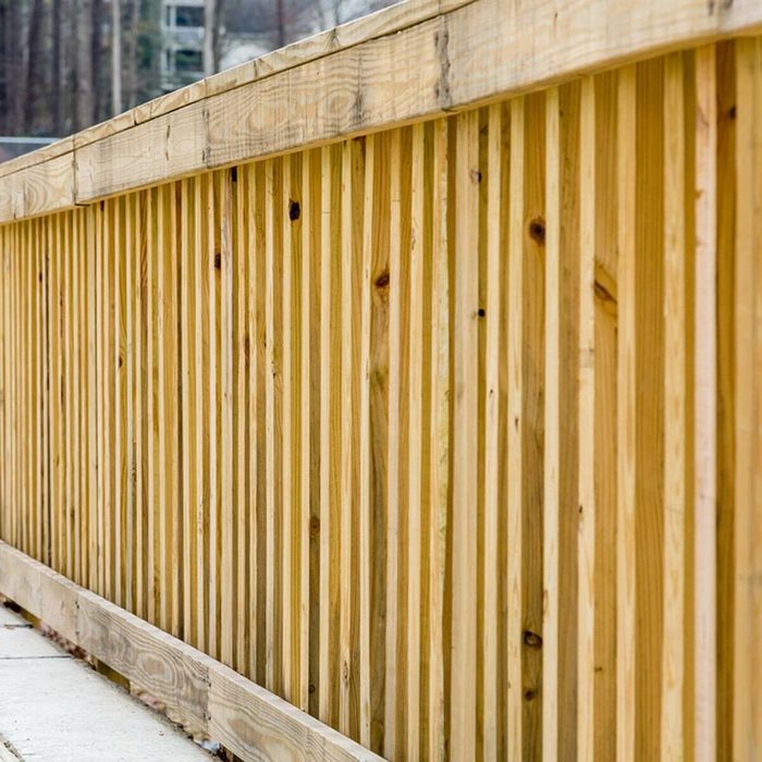 A wooden fence with vertical slats lines a concrete pathway, creating a boundary in a tranquil outdoor setting characterized by trees in the background.