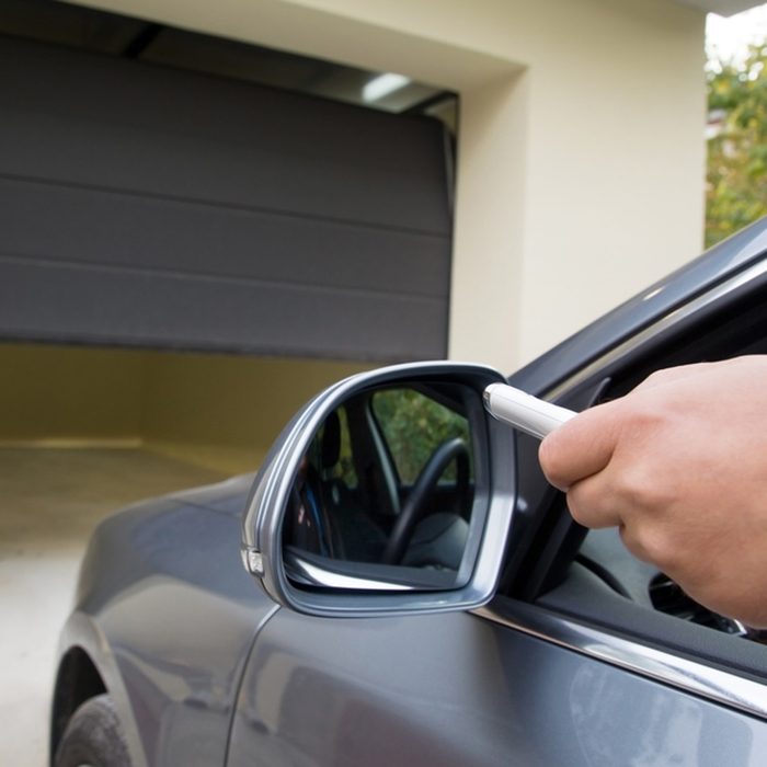 A hand holding a remote aims toward a garage door, which is partially open, as a gray car is parked nearby in a residential setting.