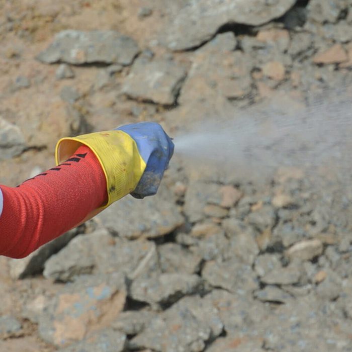 A gloved hand sprays water over a rough terrain, likely to suppress dust or assist with demolition work, surrounded by scattered rocks and dirt.