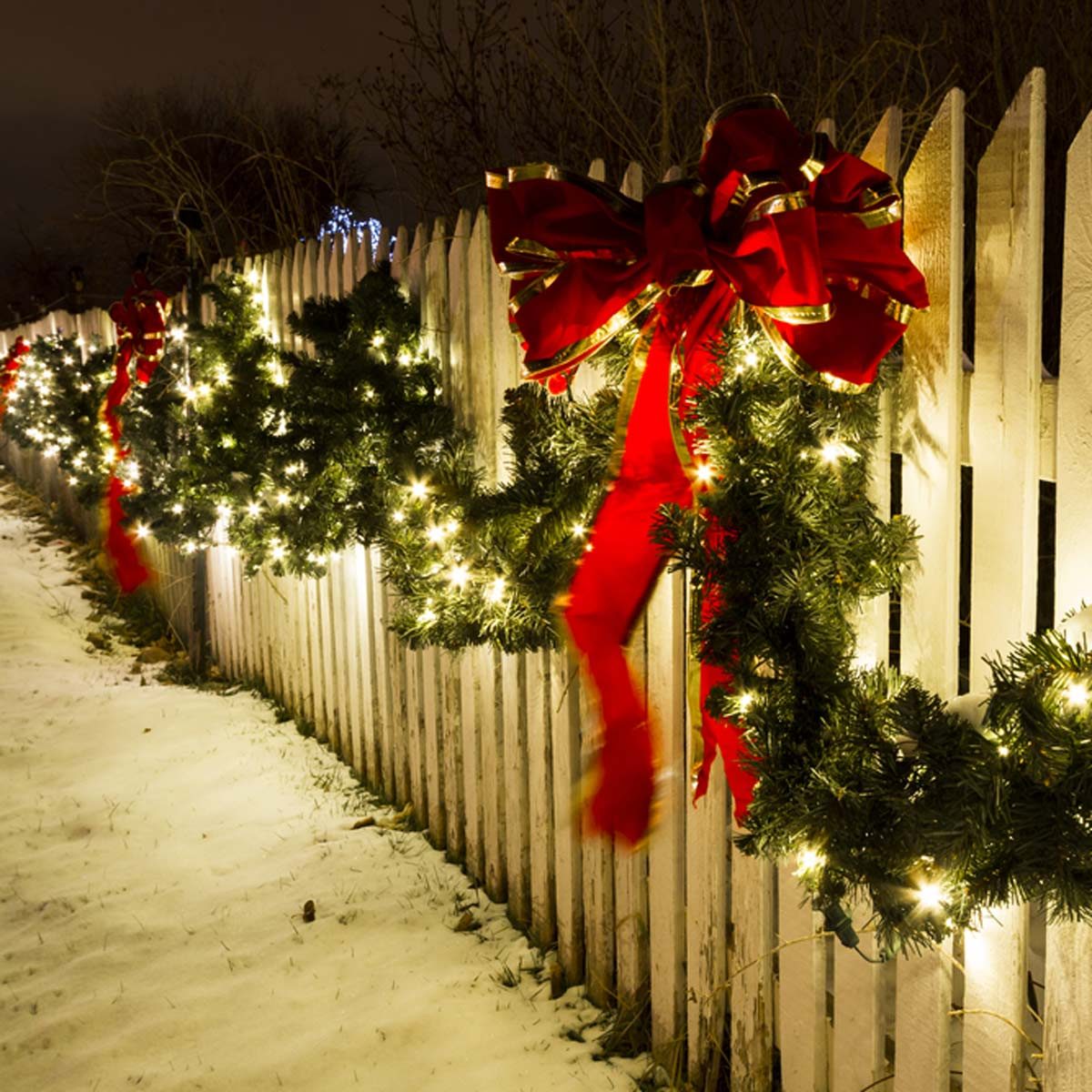 Green wreaths adorned with glowing lights and red ribbons are hung along a white picket fence, surrounded by a snowy ground at night.