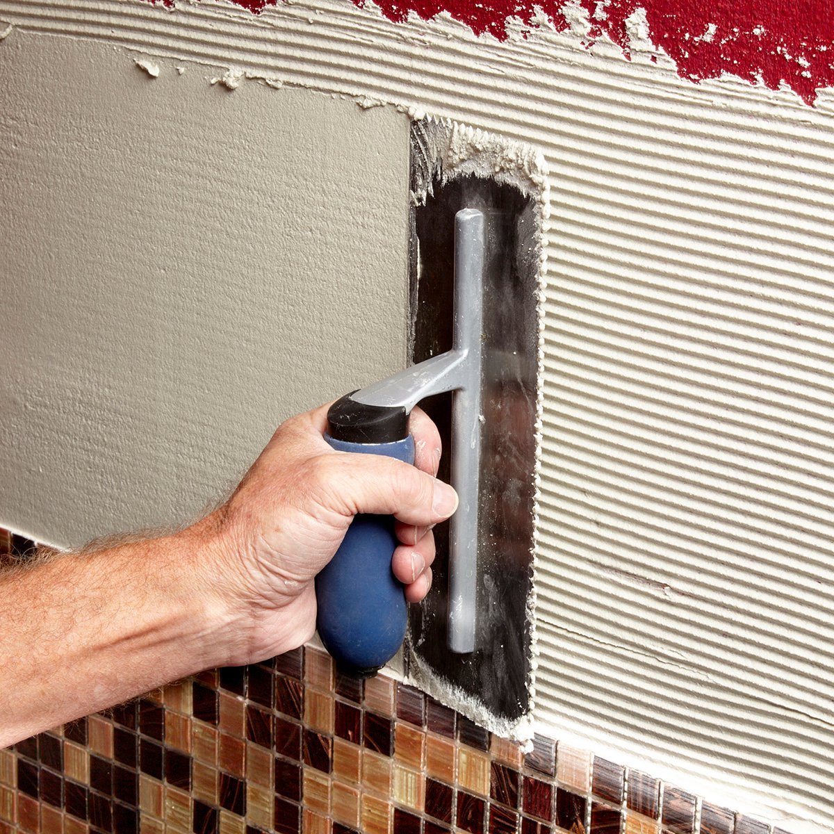 A hand holds a trowel, spreading gray material on a wall with a textured surface, near a tiled border at the bottom.