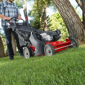 A person operates a red lawn mower, trimming grass in a green outdoor area surrounded by trees.