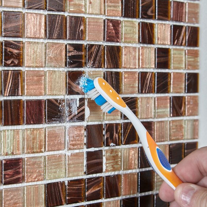 A toothbrush brushes against a textured mosaic tile wall, removing residue. The environment is indoors, emphasizing the cleaning action on the colorful tiles.
