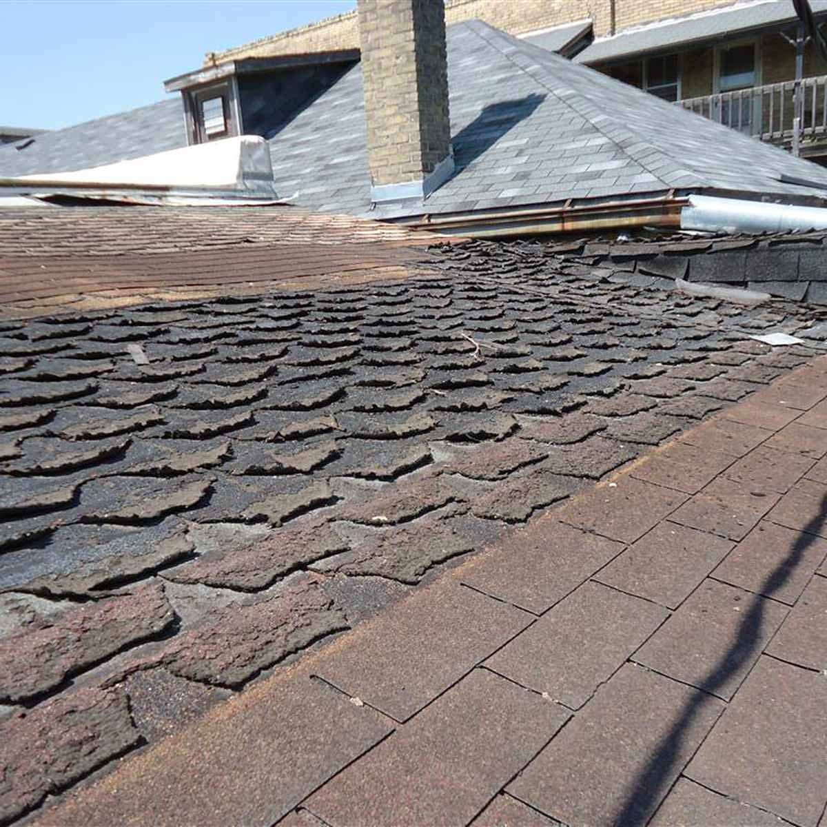 A weathered roof shows uneven shingles, with a visible chimney and adjacent sloped roof sections, under a clear blue sky.