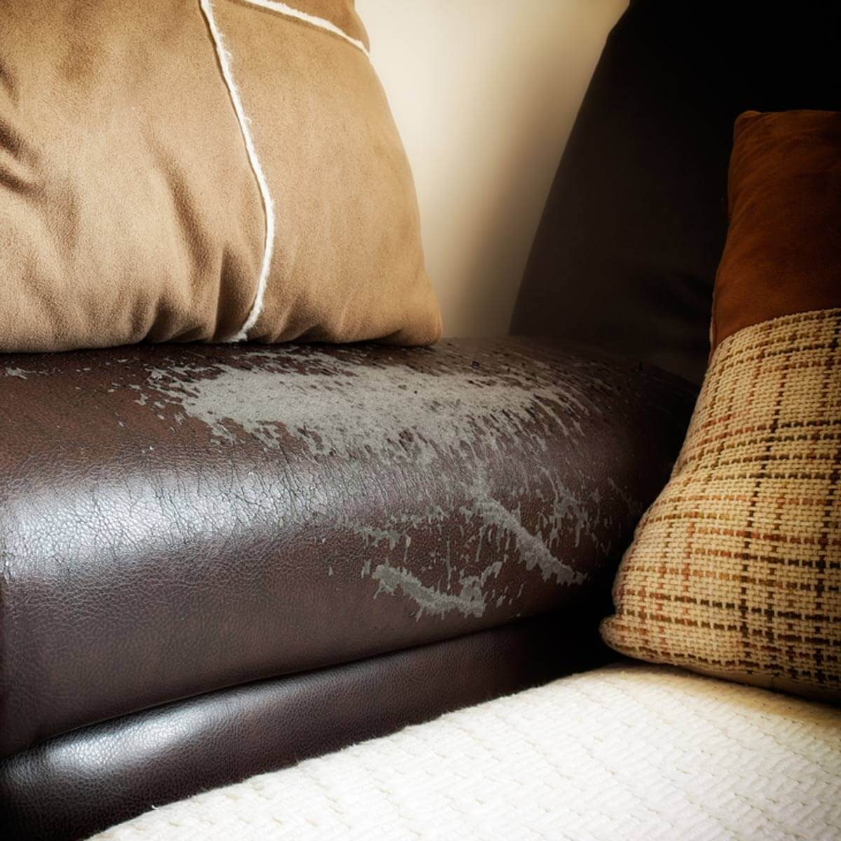A brown leather couch is stained with gray water marks, surrounded by various decorative pillows in different textures and colors.