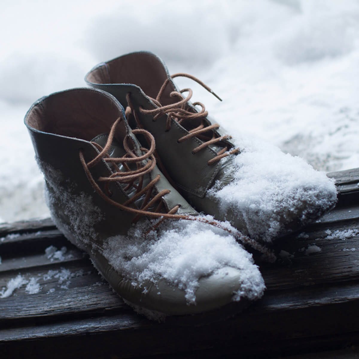 Footwear covered in snow sits on a wooden surface, indicating recent activity in a cold, wintry environment. Snow drifts are blurred in the background.
