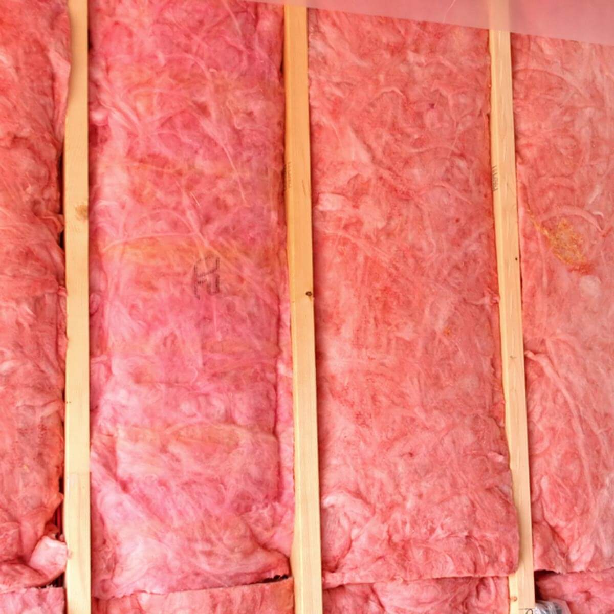 Pink insulation material fills spaces between wooden studs in a wall, providing thermal insulation in an unfinished or under-construction area.