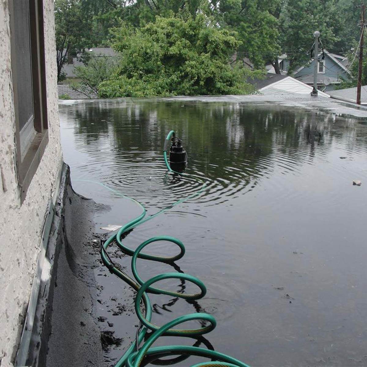 A black pump is positioned on a flooded rooftop, draining water through a green hose, surrounded by trees and nearby houses in the background.