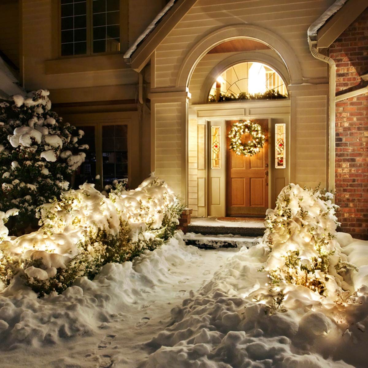 A warmly lit entrance features a door adorned with a wreath, surrounded by snow-covered plants and twinkling lights, creating a cozy winter scene.