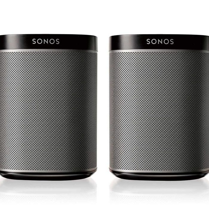 Two cylindrical speakers are positioned side by side, displaying a sleek, black finish with perforated grilles, against a clean white background.