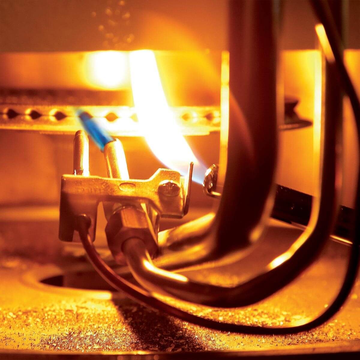A gas burner emits a blue flame, surrounded by metallic components and tubing, with an orange glow illuminating the scene.