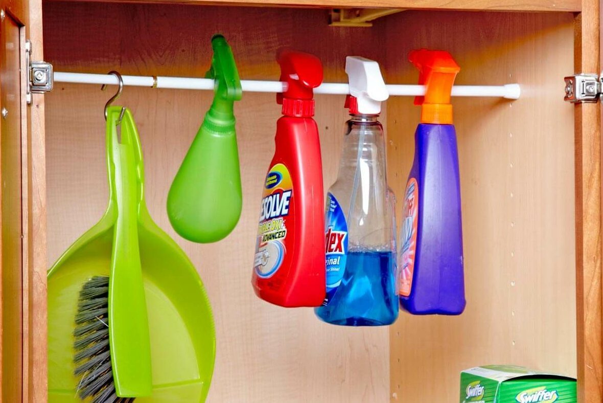 Cleaning supplies hang from a rod inside a cabinet, including spray bottles and a dustpan, with sponges and Swiffer products on the shelf below.