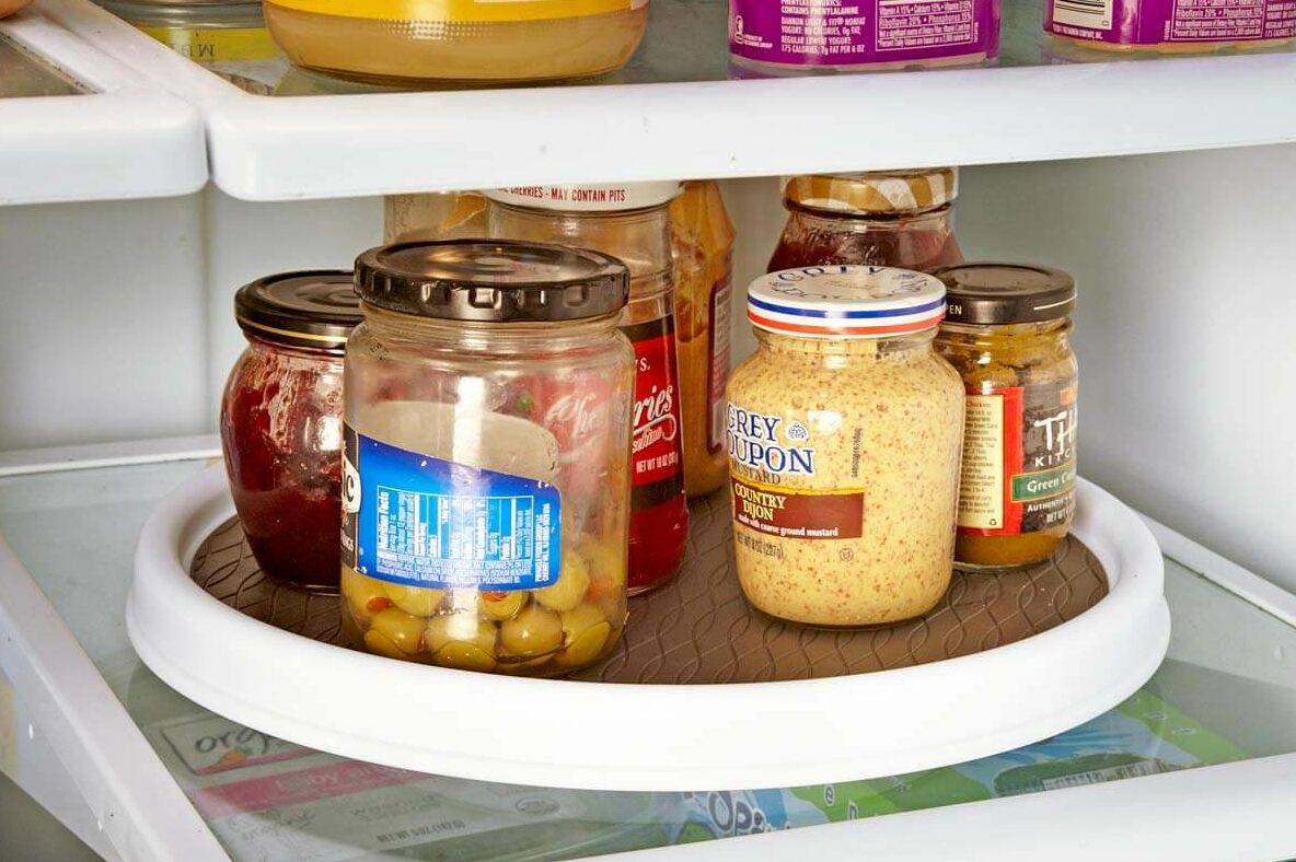 A rotating tray holds assorted jars, including mustard and olives, inside a refrigerator filled with various condiments and food items.