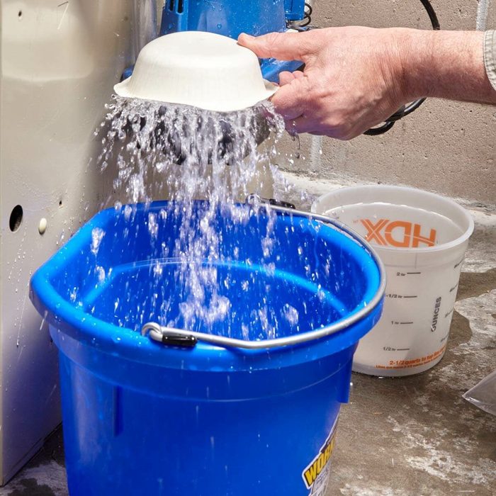 A hand tilts a white bowl, pouring water into a blue bucket below, surrounded by a concrete floor and a measuring cup nearby.