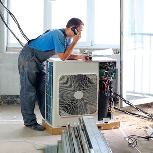 A technician is on the phone while leaning over an air conditioning unit in a partially renovated room, surrounded by construction materials and unfinished walls.
