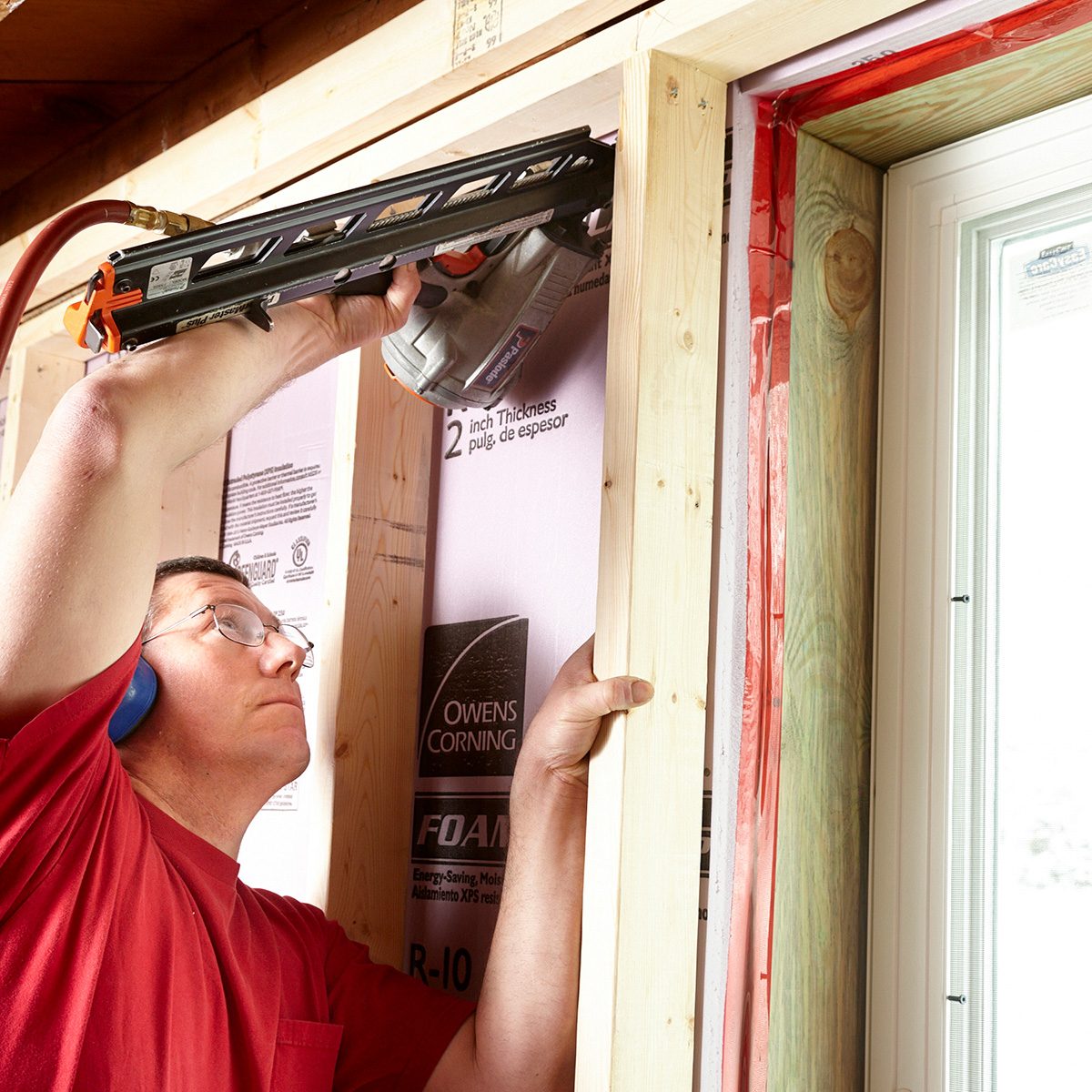 A man uses a power tool to secure a wooden frame around a window. Insulation is visible between the frame and the wall.