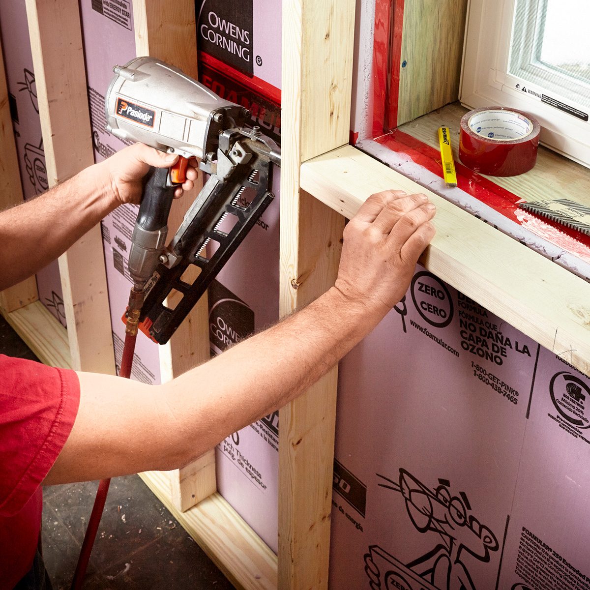 A person uses a pneumatic nail gun to attach wooden framing in a construction setting, with insulated wall panels and tools nearby.
