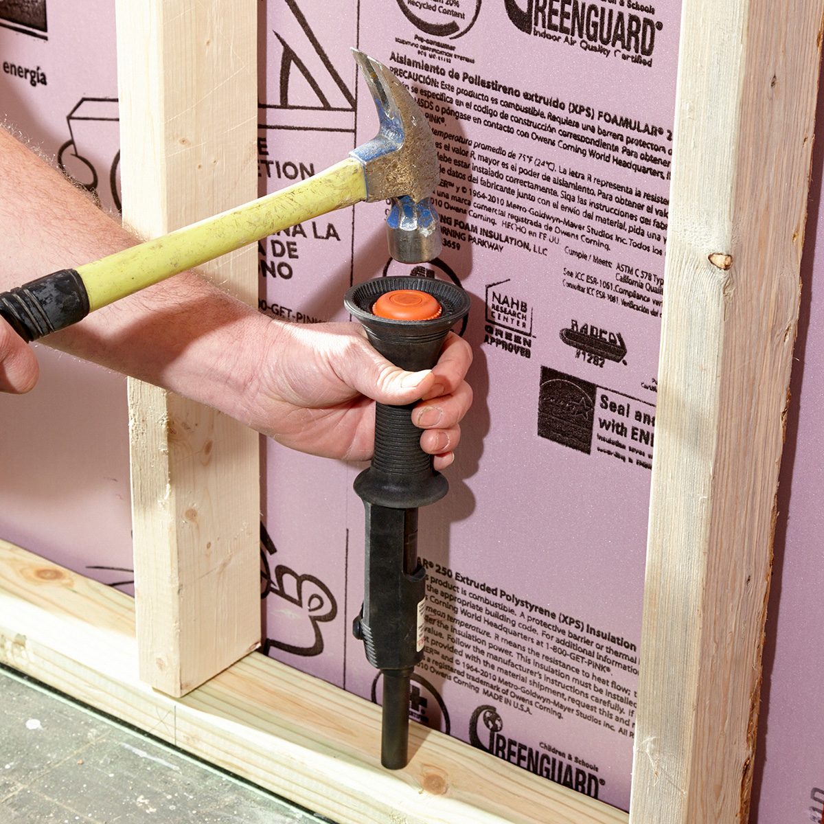 A hand holds a black tool against a pink insulation board while a hammer strikes the tool, set within a wooden frame structure.