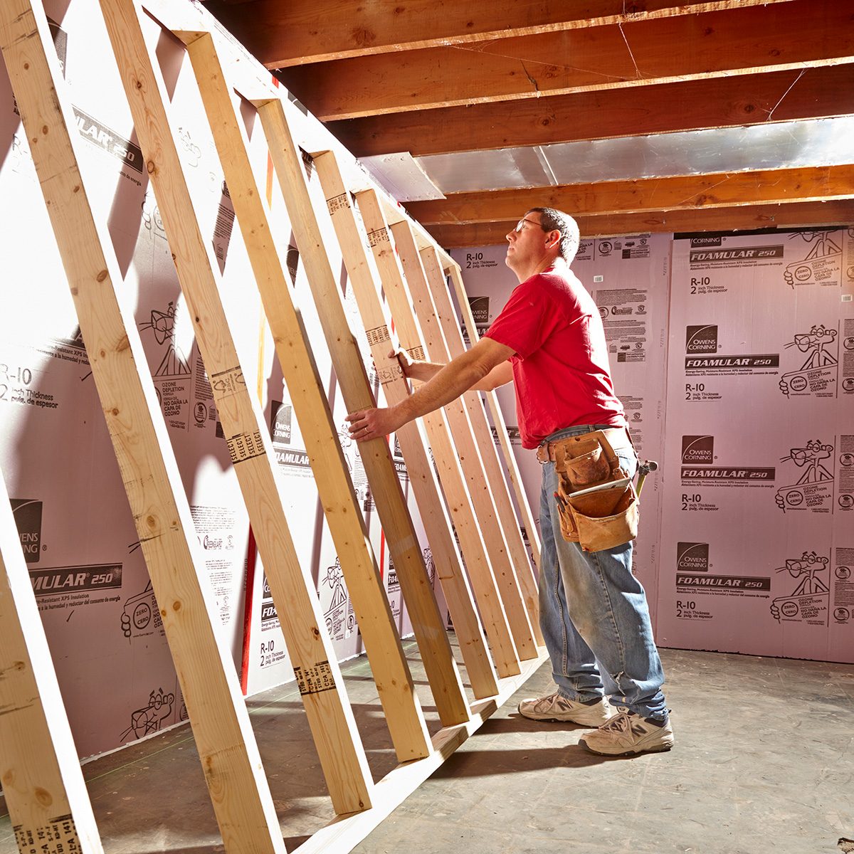 A man adjusts wooden framing in a partially finished room, surrounded by insulation on the walls and wooden beams overhead.