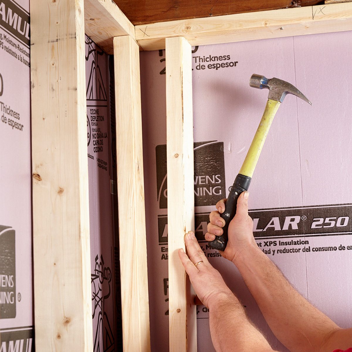 A hand holds a hammer, striking a wooden stud against a pink insulation background, engaged in a home construction task within a framed structure.