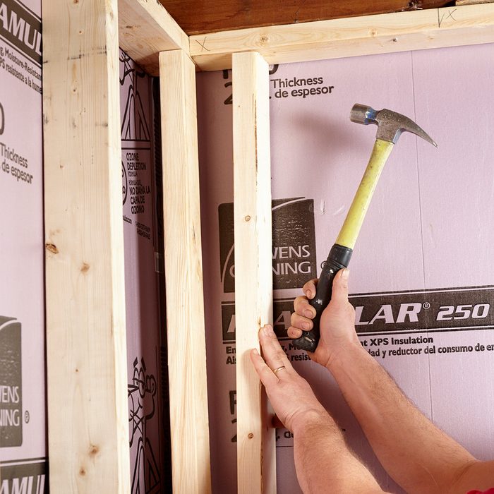 A hand holds a hammer, striking a wooden stud against a pink insulation background, engaged in a home construction task within a framed structure.