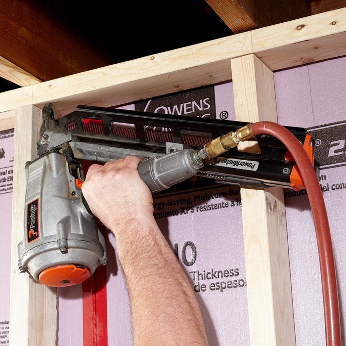 A person uses a nail gun to attach framing to a wall, surrounded by insulation board and wooden structures in a construction setting.