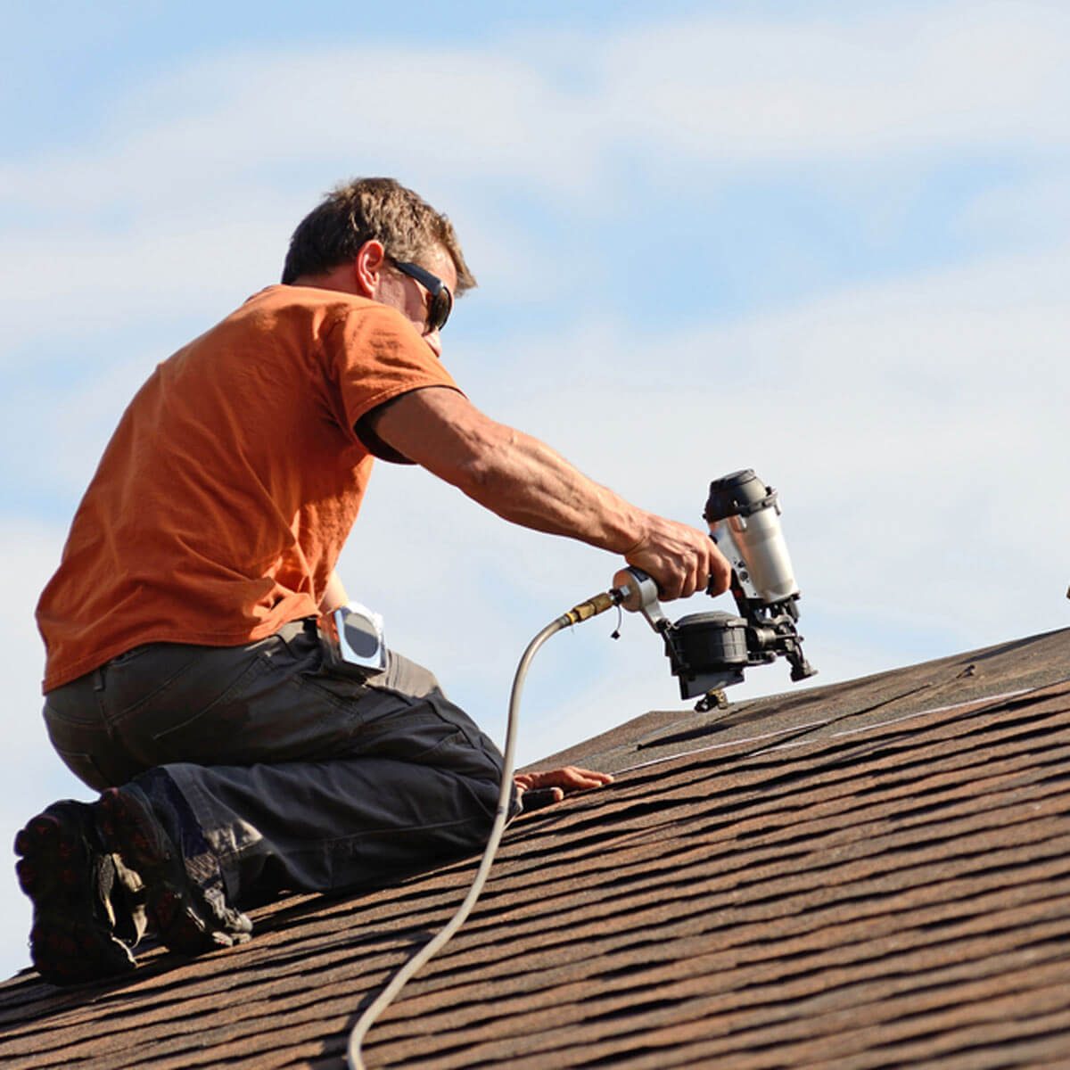 A man in an orange shirt uses a nail gun on a sloped roof, surrounded by blue sky and clouds.