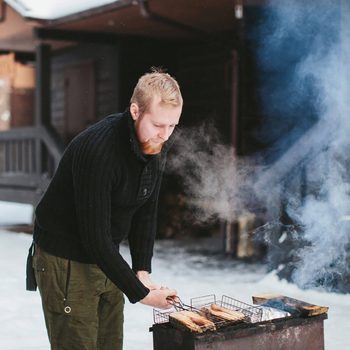 A man grills food over a smoky fire pit while surrounded by snow, wearing a black sweater and green pants, concentrating on his task.