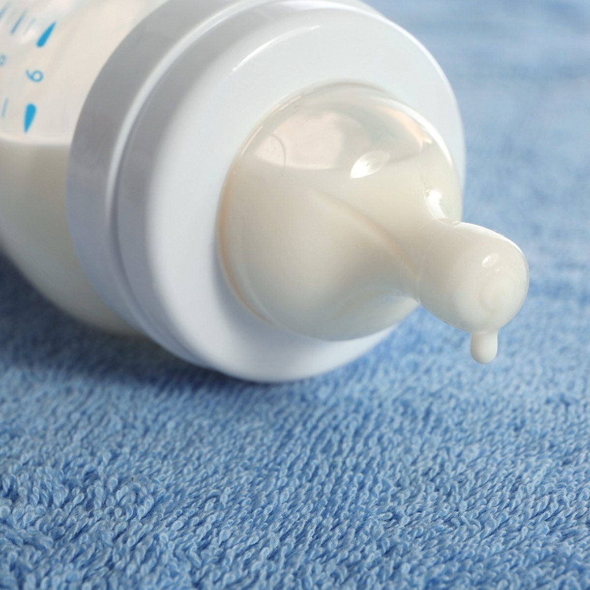 A baby bottle, slightly tipped, drips white liquid onto a soft blue towel, indicating recent use or preparation for feeding.
