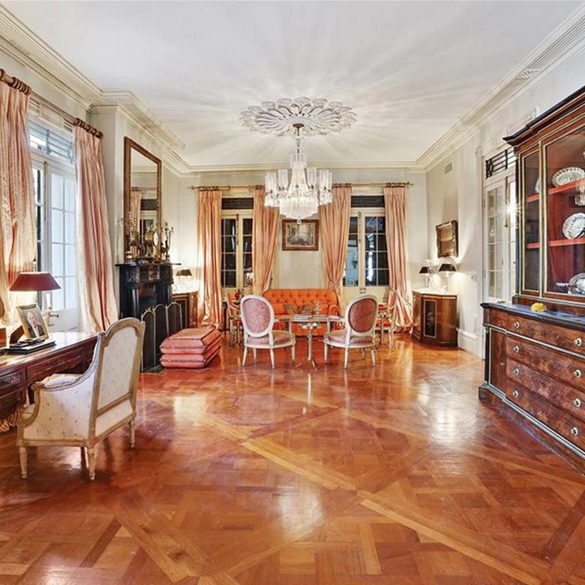 A stylish living room features an orange sofa, glass chandelier, and ornate furniture. Large windows let in natural light, enhancing the elegant ambiance.