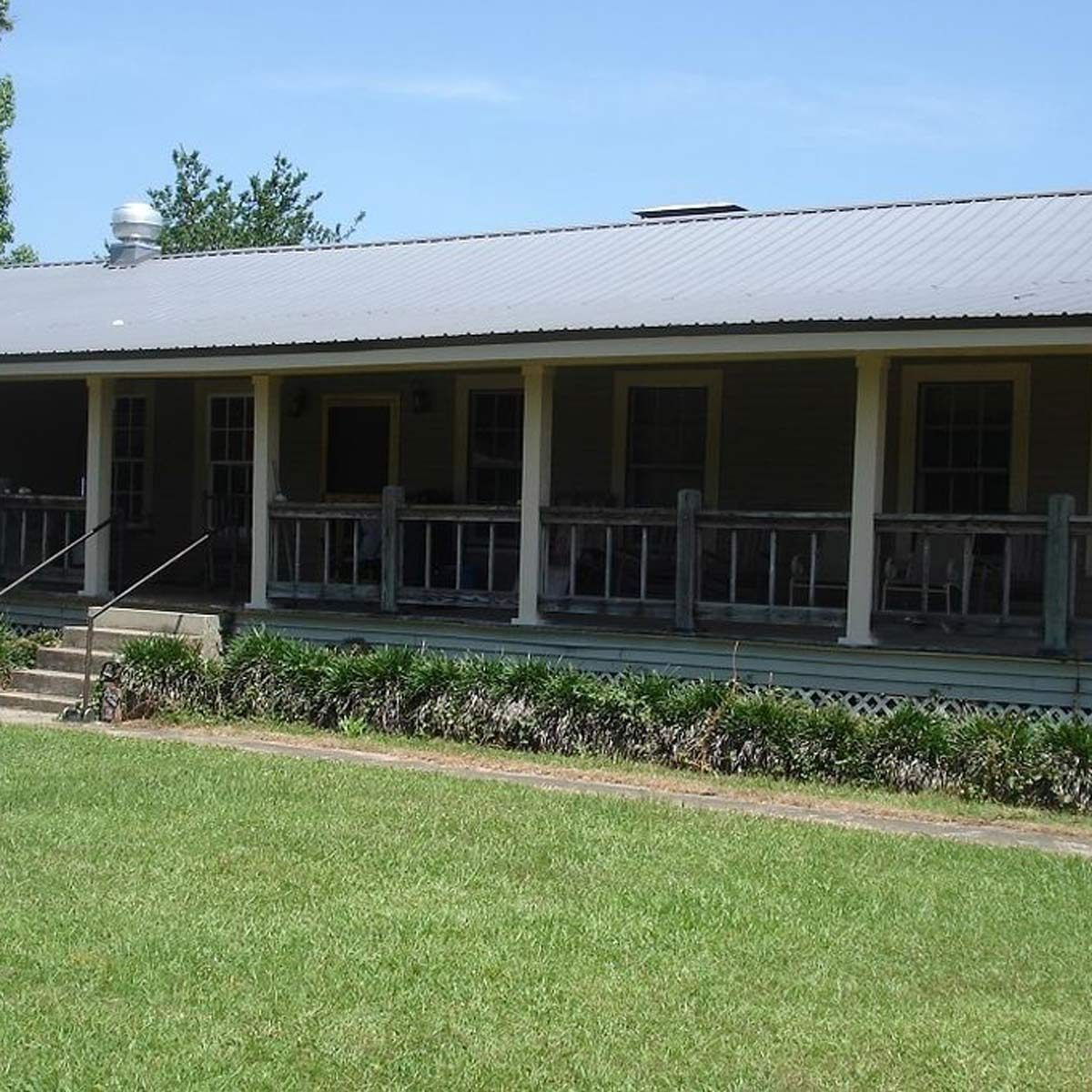 A wooden house with a wide porch, steps leading to a grassy yard, surrounded by shrubs under a clear blue sky.