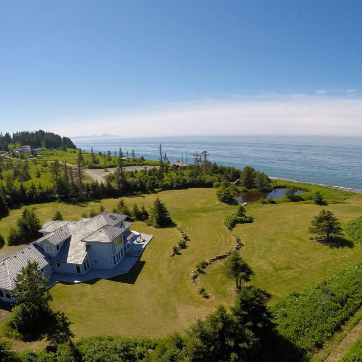 A seaside house rests in a spacious lawn, surrounded by trees and a pond, under a clear blue sky, with ocean waves in the background.