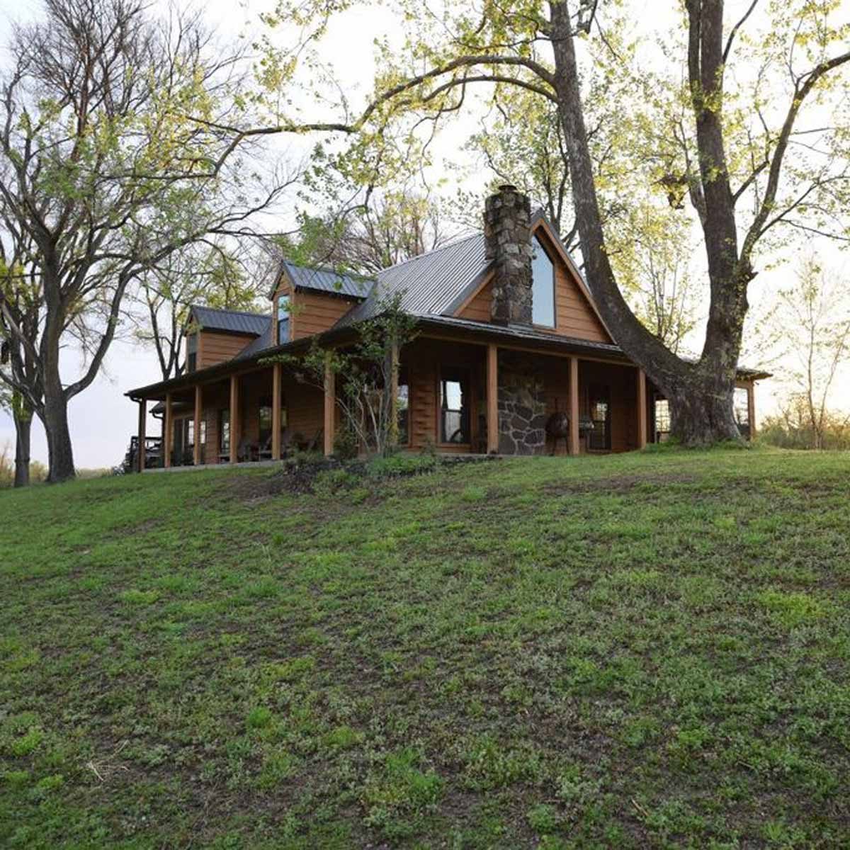 A rustic wooden house with a stone chimney stands on a grassy slope, surrounded by trees, under a clear sky during early evening.