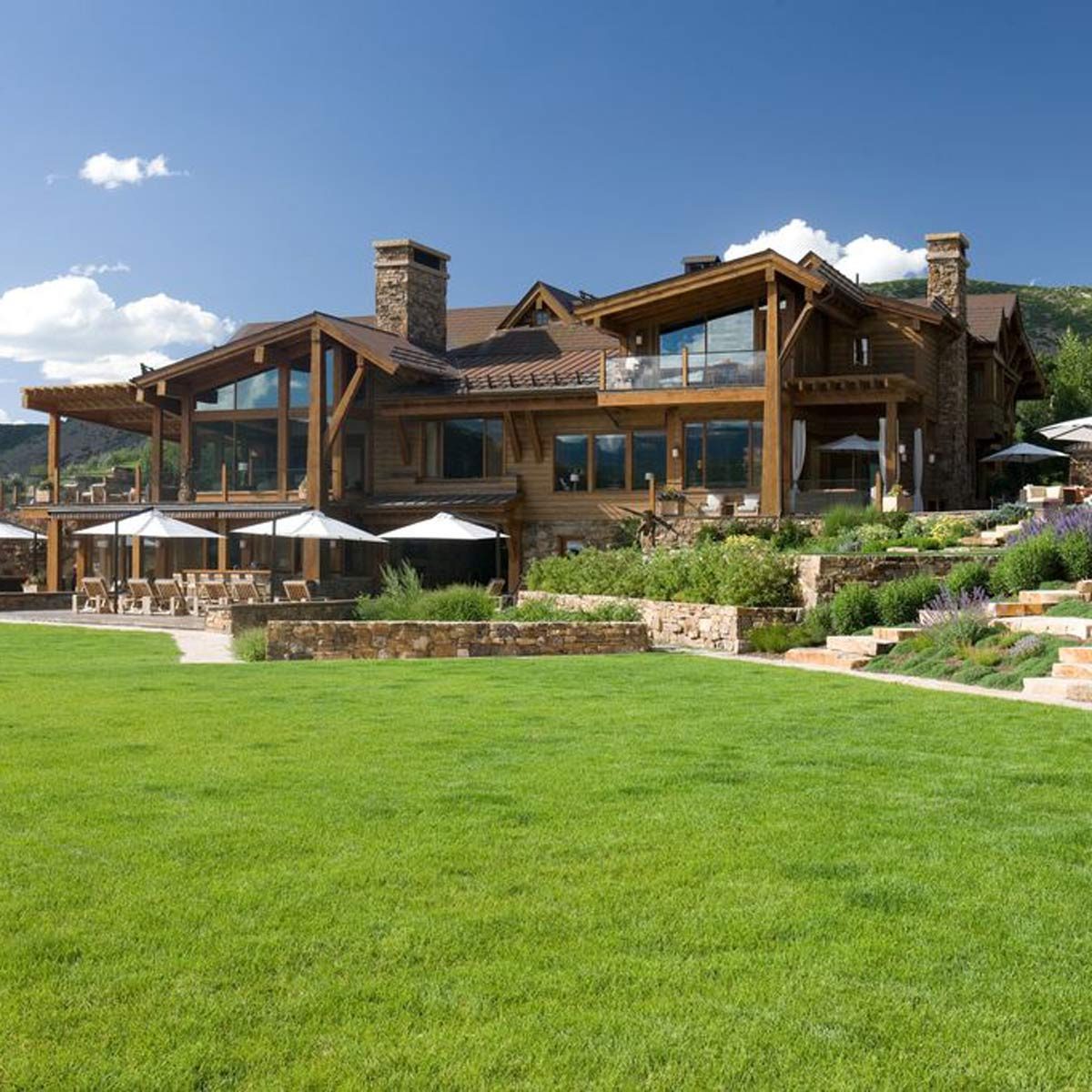 A large wooden house with expansive windows sits beside a green lawn, featuring stone landscaping and outdoor seating shaded by umbrellas under a clear blue sky.