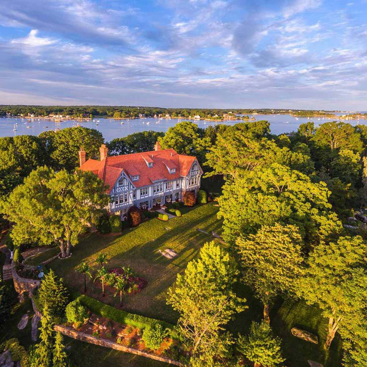 A large, elegant house with a red roof sits amidst lush trees, overlooking a calm body of water with boats, under a partly cloudy sky.