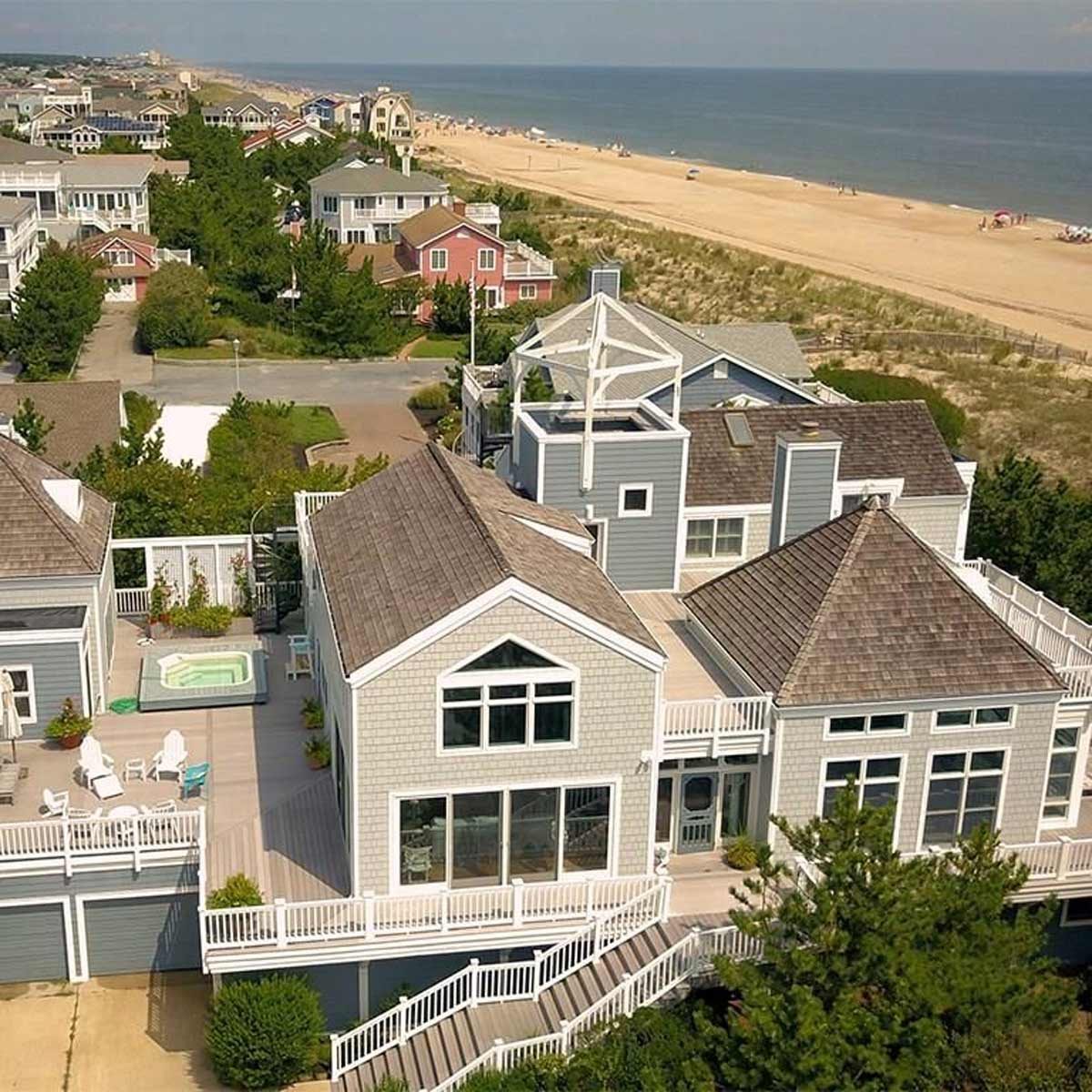 A large, modern beach house with multiple roofs and a hot tub is situated near a sandy beach, surrounded by other residential buildings and greenery.