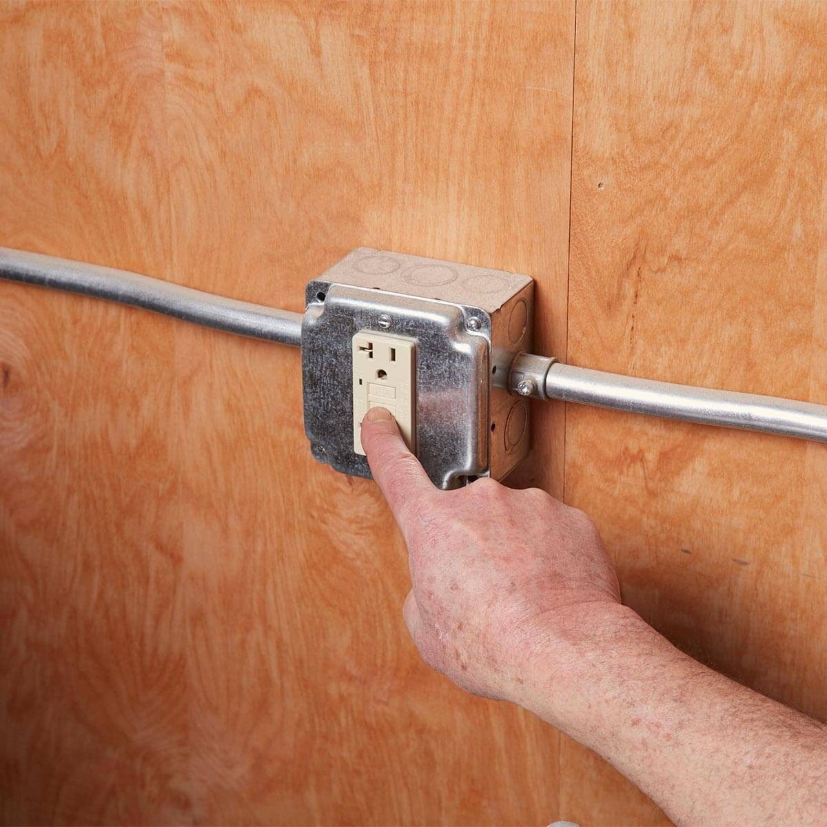 A hand presses a power outlet on a wooden wall, with electrical conduit visible. The outlet is mounted in a metal box.