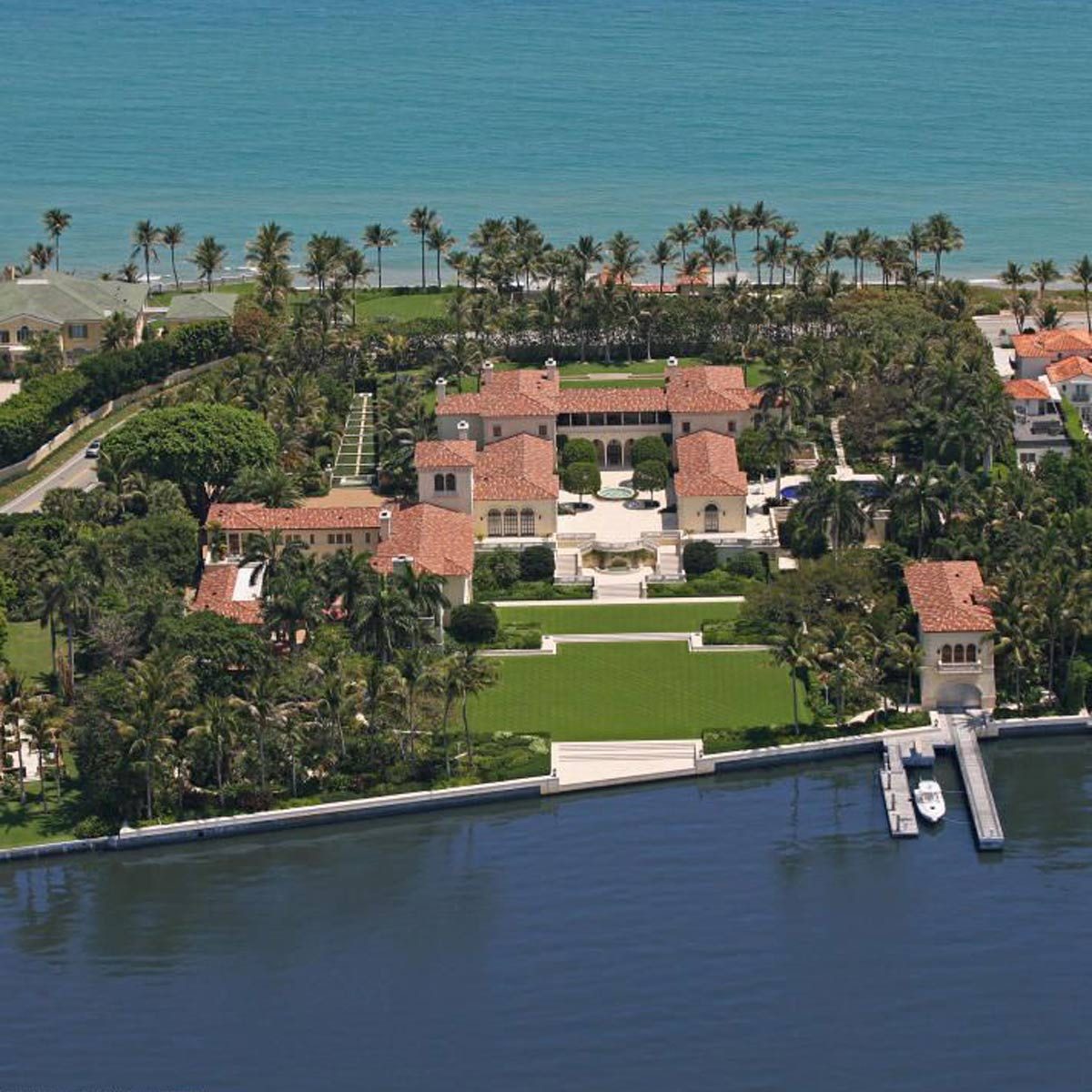 A luxurious estate features multiple buildings with terracotta roofs, surrounded by vibrant greenery and palm trees, adjacent to a calm blue waterfront.