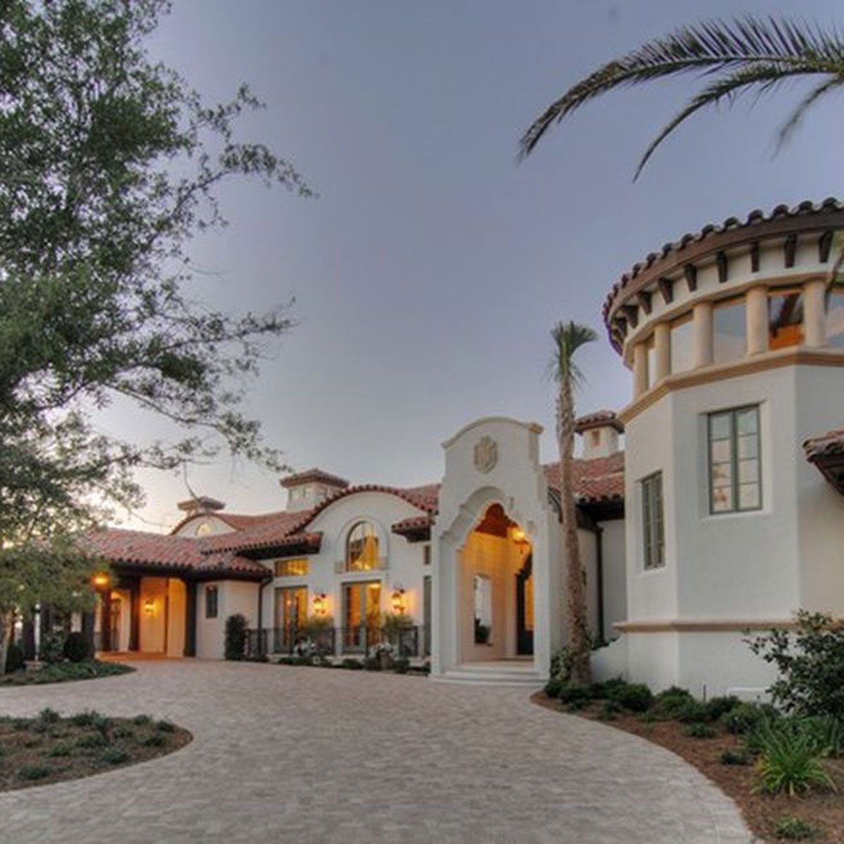 A large, elegantly designed house sits at twilight, featuring arched windows and a circular turret, surrounded by trees and manicured landscaping along a paved pathway.