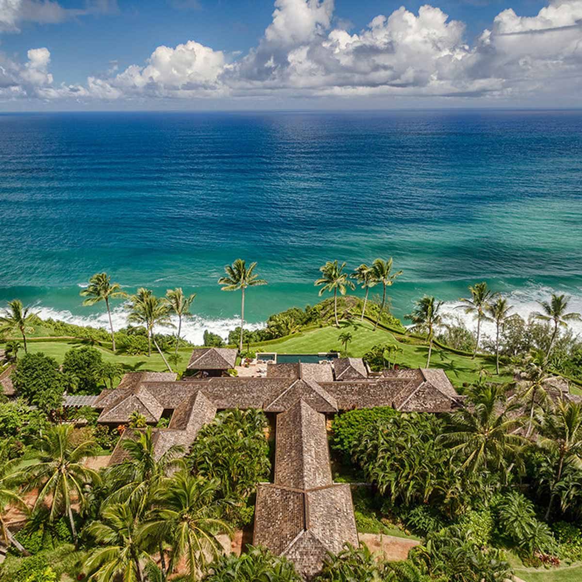 A cluster of tropical homes rests amid lush greenery, while waves crash against a nearby shoreline under a vibrant sky filled with clouds.